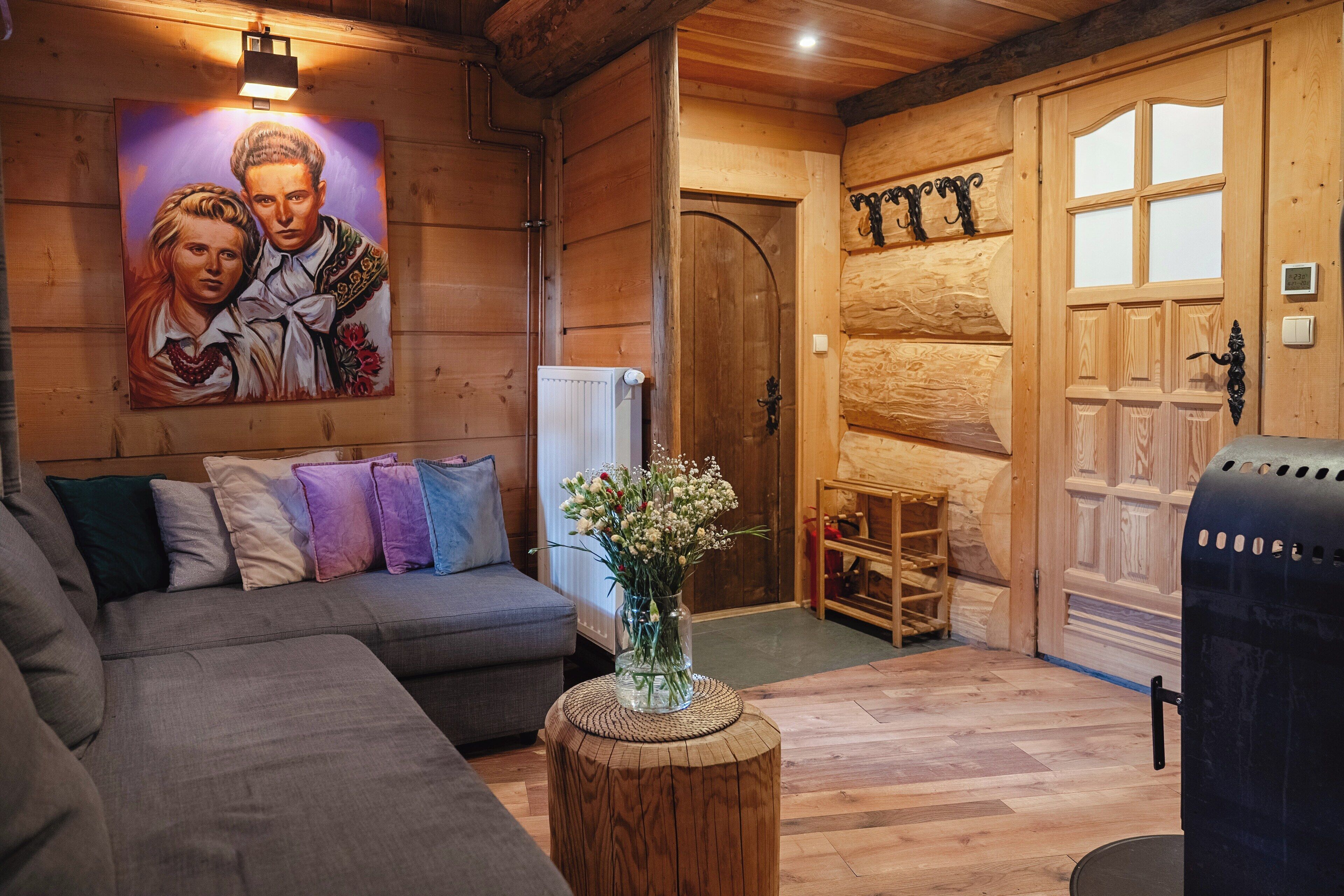 A cozy log cabin interior with a sofa, a wooden coffee table with a vase of flowers, a fireplace, and a door with decorative elements.