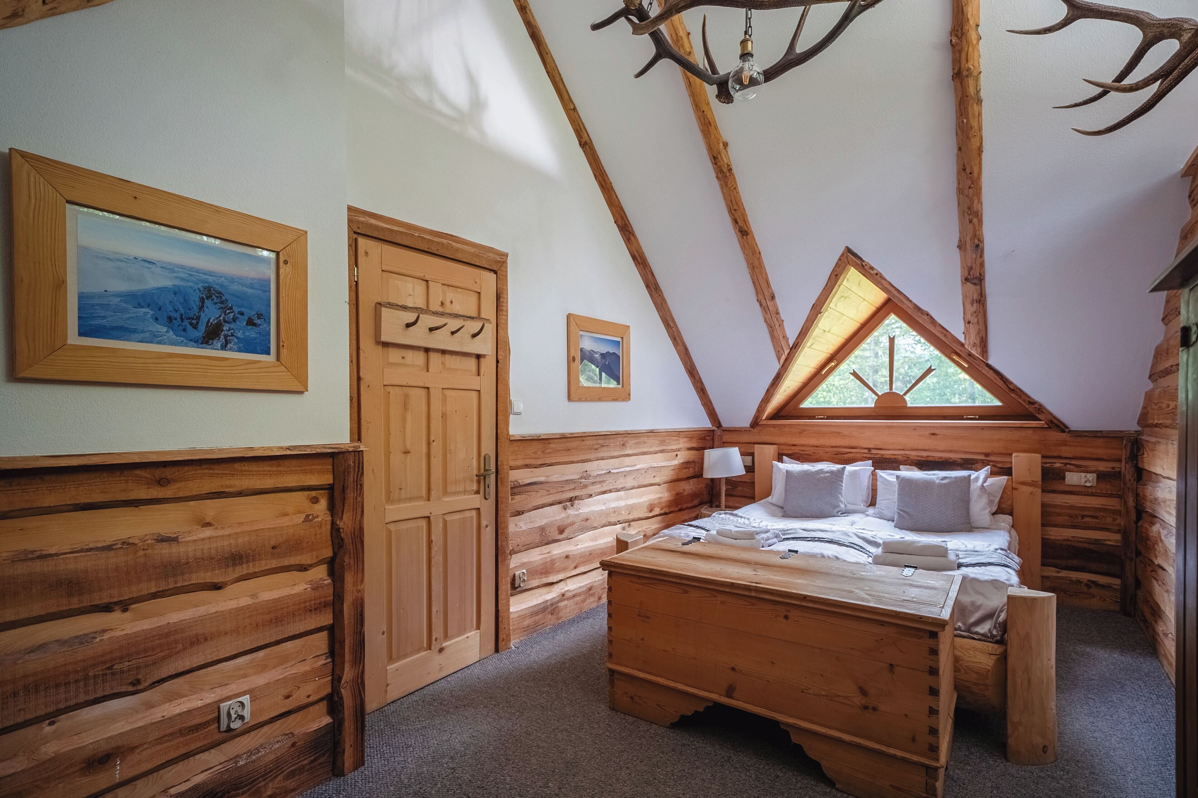 A log cabin bedroom with a bed, a wooden dresser, a framed picture, a door, and a window with a view of trees.