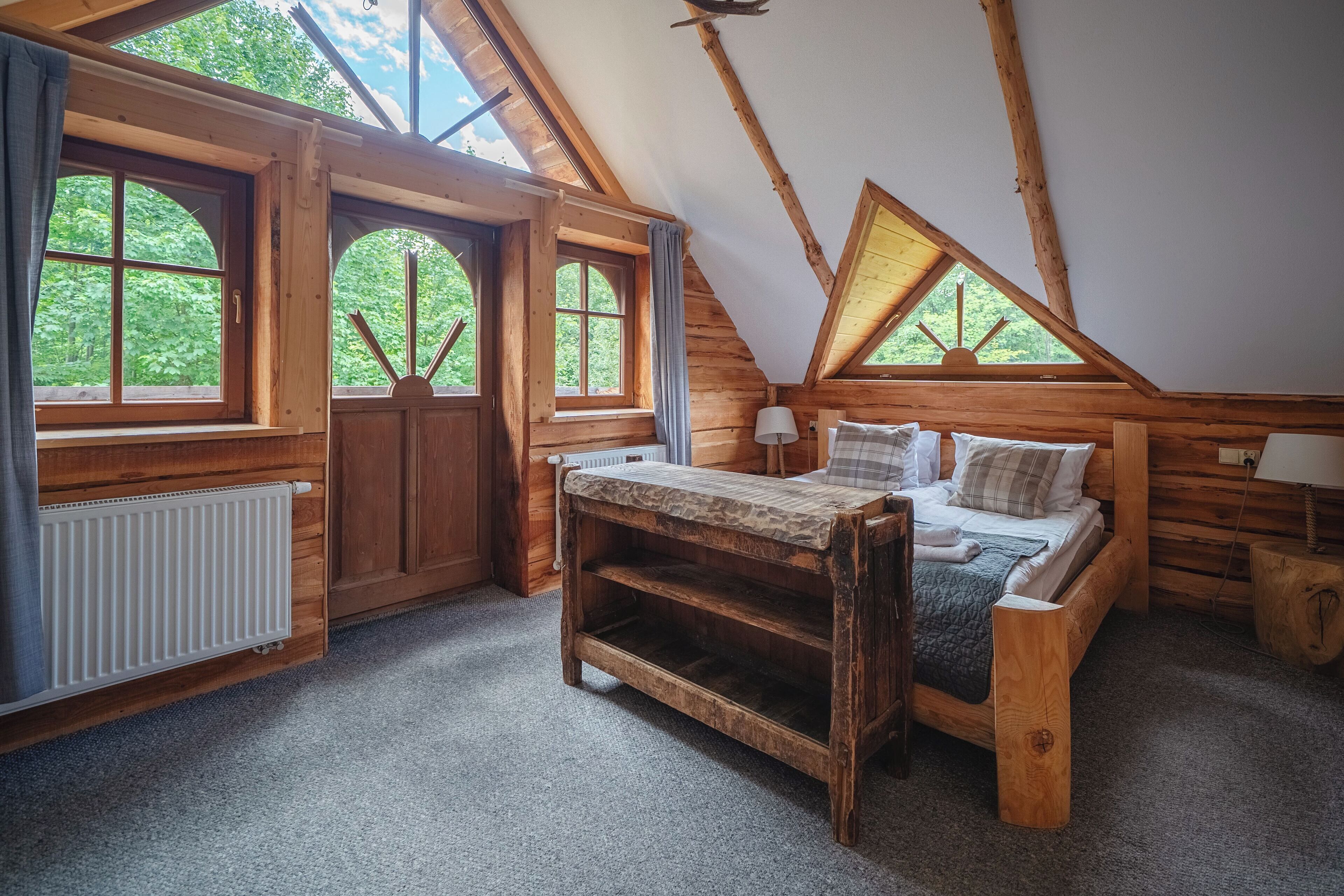 Une chambre mansardée chaleureuse, avec un lit en bois, une petite table et de grandes fenêtres donnant sur la verdure environnante.