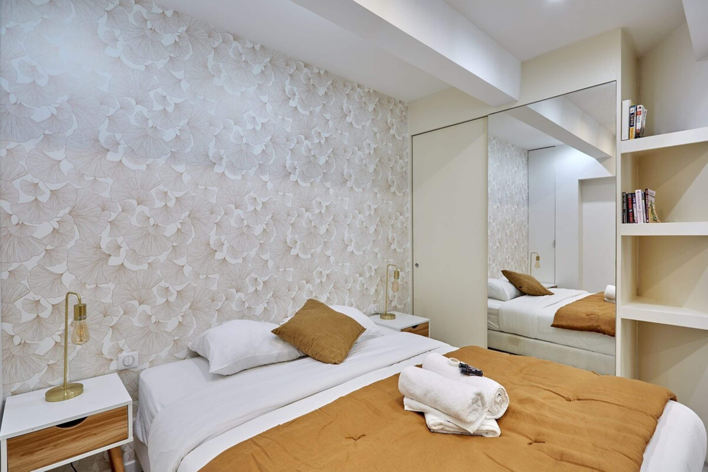 A modern hotel room with a large bed, a nightstand, a mirror, and a bookshelf.