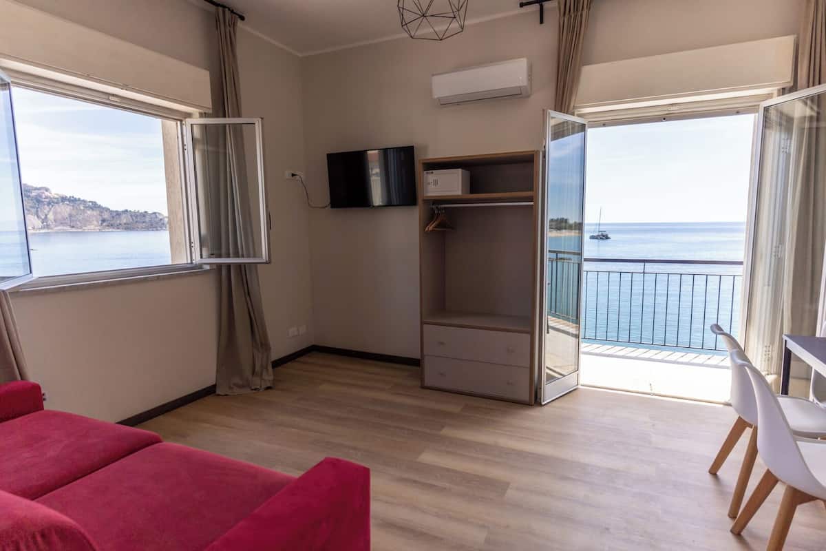 Ein Zimmer mit großem Fenster, Fernseher, Kleiderschrank und einem Balkon mit Meerblick.