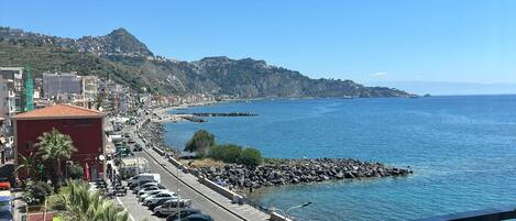 Un balcón con mesa y sillas que da a un pueblo costero, con vistas claras al mar y una colina distante.