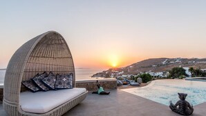 Una silla de mimbre con cojines, una pequeña estatua y vistas a la piscina con un atardecer sobre un pueblo costero.