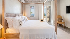 Una cama con dosel, ropa de cama blanca, almohadas con estampado de estrellas, cabecera de madera y vista al mar a través de una ventana.