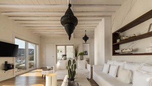 Amplia sala de estar con sofá blanco, una mesa de centro y un cactus como elemento decorativo. Se observan dos faroles colgantes, un televisor de pantalla plana montado en la pared y vistas al exterior a través de amplias ventanas.
