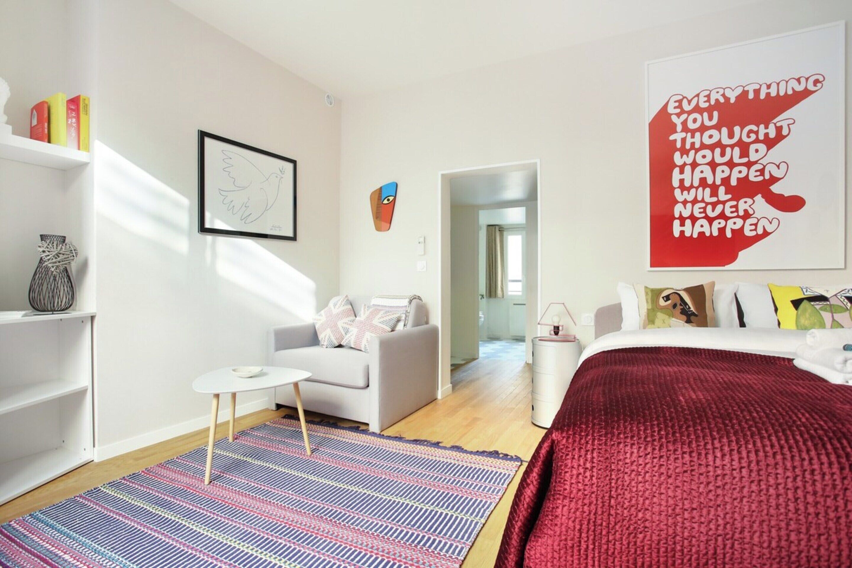 A modern hotel room with a bed, a sofa, a small table, and a colorful rug. There are books on a shelf, a framed artwork, and a motivational quote on the wall.