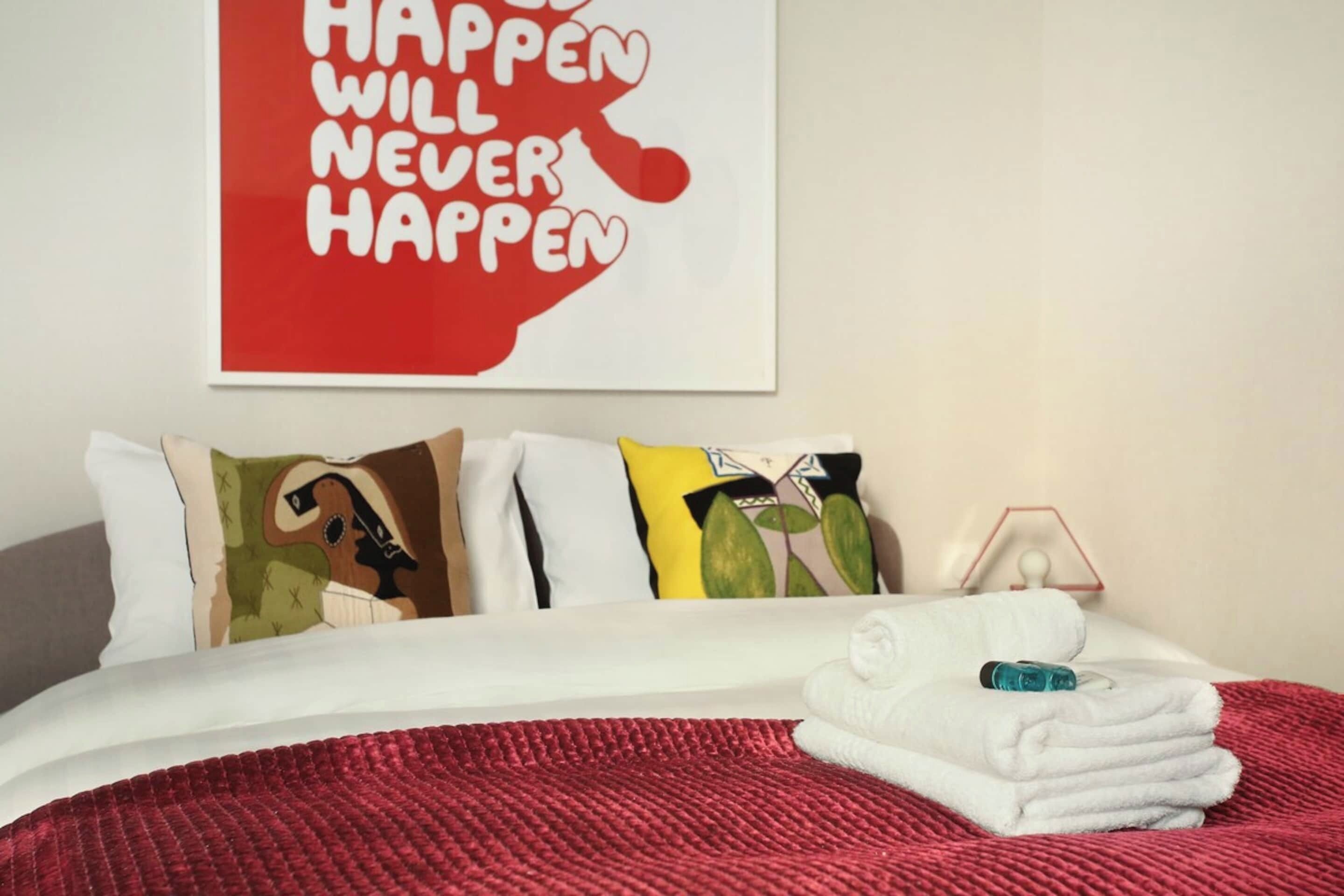 A neatly made bed with white linens, a red blanket, and a folded towel. A colorful pillow with a leaf design is visible. A red poster with white text is mounted on the wall above the bed.