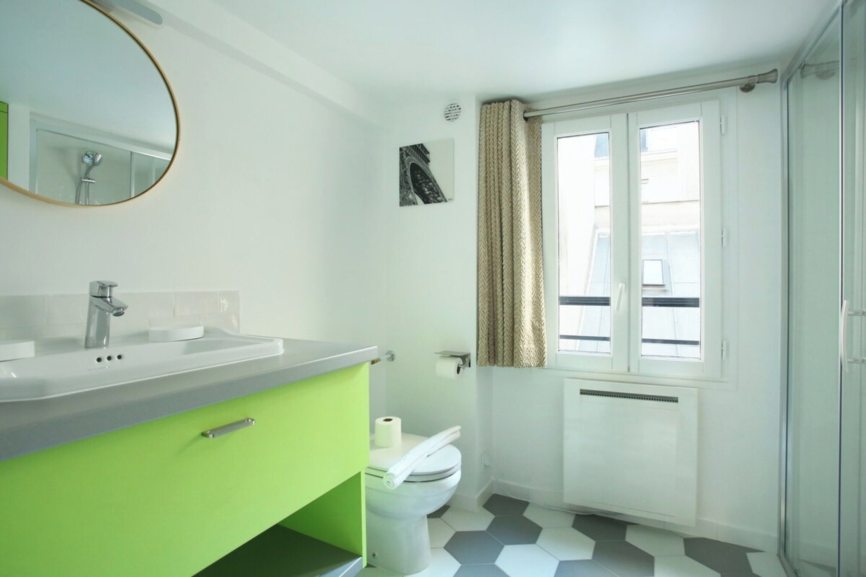 Une salle de bain avec un lavabo blanc, un meuble vert, des toilettes et une fenêtre avec des rideaux.