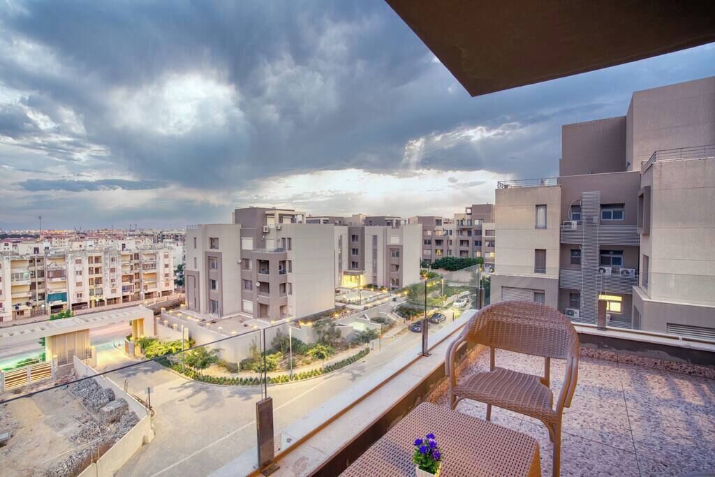 A balcony with a wicker chair and a table, overlooking a residential area with modern buildings and a road.