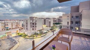 A balcony with a wicker chair and a table, overlooking a residential area with modern buildings and a road. - The Suite, Palm Hills - VG631, Residential Suites near AUC by ELEGANT (New Cairo City)