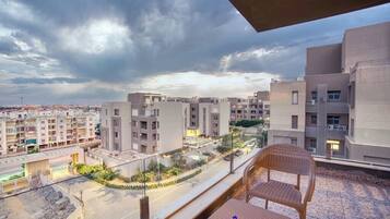 A balcony with a wicker chair and a table, overlooking a residential area with modern buildings and a road.