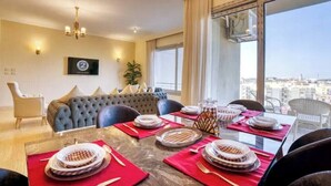 A dining area with a large table set for four, a sofa, and a view of the city. - The Suite, Palm Hills - VG631, Residential Suites near AUC by ELEGANT (New Cairo City)