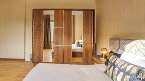A bedroom with a bed, a nightstand with a lamp, and a wooden wardrobe with a mirror. - The Suite, Palm Hills - VG631, Residential Suites near AUC by ELEGANT (New Cairo City)