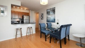 A modern kitchen with stainless steel appliances, a dining area with blue chairs and a glass table, and a large abstract painting on the wall. - High-Rise Loft & Panoramic Harbour Views (Victoria Central British Columbia Provincial Government)