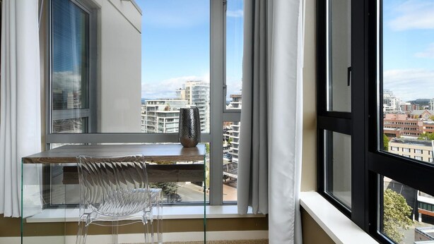 A balcony with a glass table and chair, a decorative vase, and a view of city buildings. - High-Rise Loft & Panoramic Harbour Views (Victoria Central British Columbia Provincial Government)