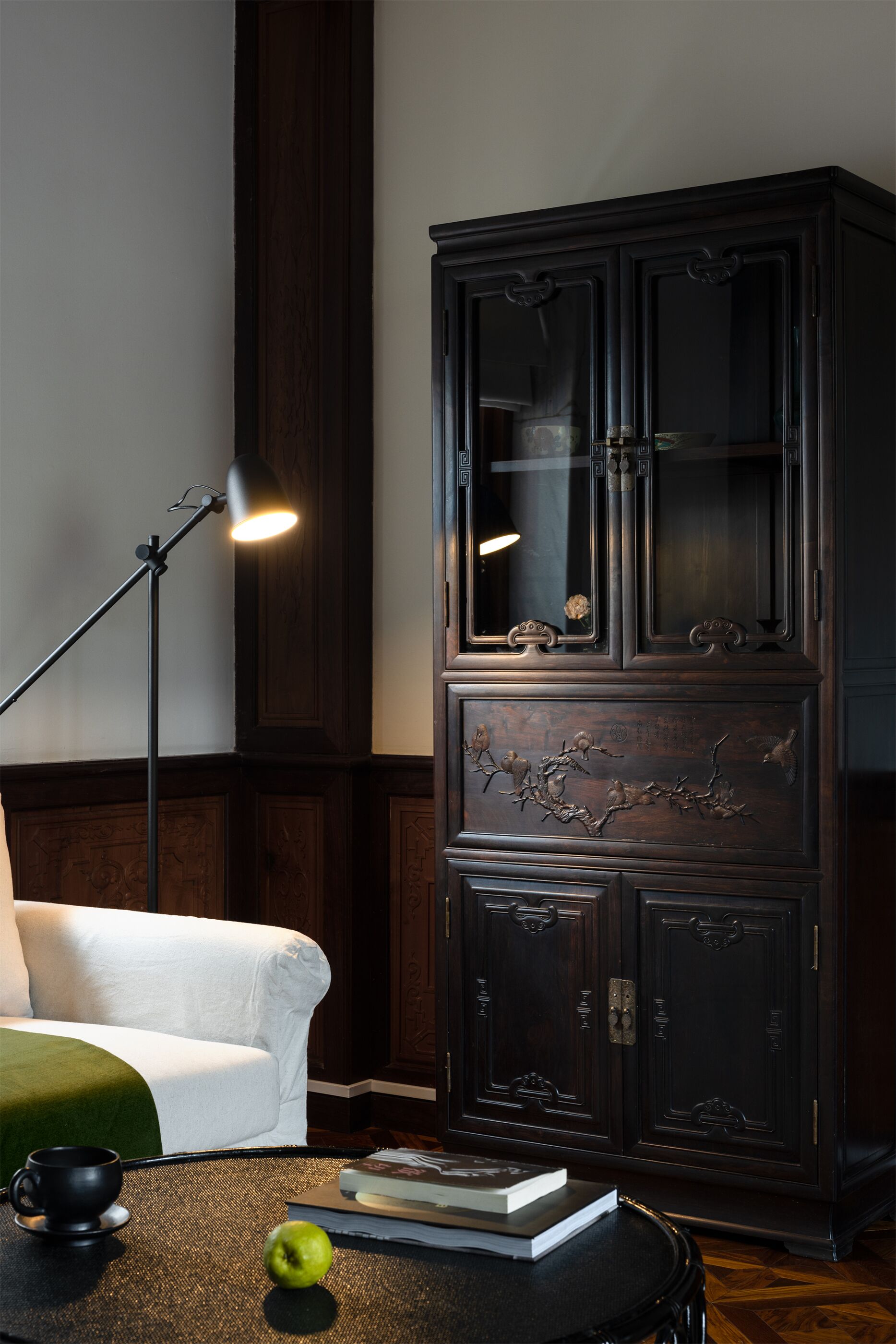 A hotel room with a dark wooden cabinet, a white sofa, a standing floor lamp, and a small round table with a book, a cup, and an apple.