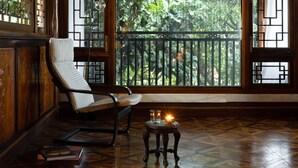 A room with a wooden floor, a cushioned chair, a small table with a candle, and a view of greenery through a window with a geometric pattern. - GRAND JOLI (Lijiang)