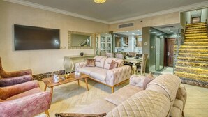 Un vaste salon comprenant un grand canapé, une table basse et un téléviseur fixé au mur. Un escalier avec une rampe dorée mène à l’étage supérieur.