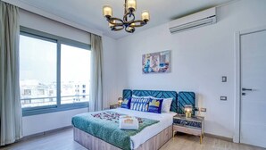 A hotel room with a large bed, a nightstand, a chandelier, a painting, and a view of the city through the window.