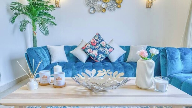 A blue sofa with floral cushions, a white vase with flowers, and a decorative metal bowl on a wooden table.