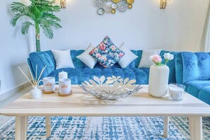 Ein blaues Sofa mit floralen Kissen, eine weiße Vase mit Blumen und eine dekorative Metallschale auf einem Holztisch.