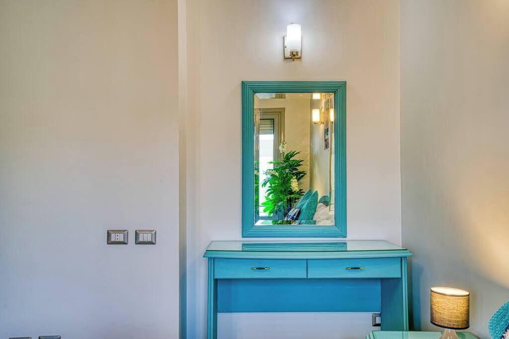 Une salle de bain avec un meuble-lavabo de couleur turquoise, un miroir, un luminaire fixé au mur et une petite table avec une lampe.