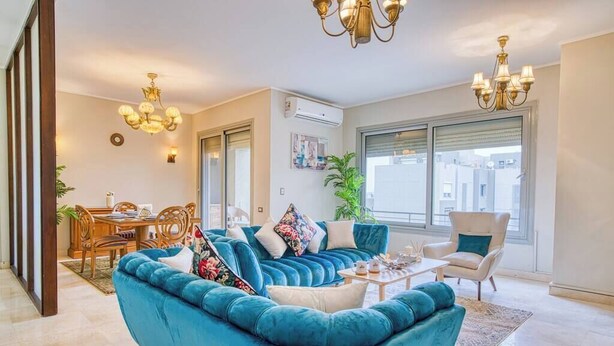 A spacious living room with a teal sofa, a dining area with wooden furniture, and a large window offering a city view. - The Suite, Palm Hills - VG924, Residential Suites near AUC by ELEGANT (New Cairo City)