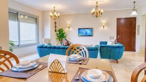 A dining area with a wooden table set for four, blue upholstered sofas, and a TV mounted on the wall. - The Suite, Palm Hills - VG924, Residential Suites near AUC by ELEGANT (New Cairo City)