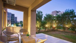 An outdoor patio area with wicker furniture and a small table, surrounded by a garden and a view of residential buildings in the background.