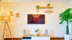 Een woonkamer met een flatscreen televisie aan de muur, een witte tv-meubel met decoratieve objecten, een vloerlamp en een grote potplant.