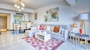 Ein modernes Wohnzimmer mit einem grauen Sofa, einem weißen Couchtisch und einem geometrischen Teppich. An den Wänden befinden sich gerahmte Kunstwerke und es gibt einen Essbereich mit einem weißen Tisch und Stühlen.