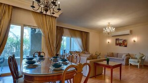 A dining area with a glass table, wooden chairs, and a chandelier. There is a sofa set and a coffee table in the living area. - The Suite, Palm Hills - V1811, Residential Suites near AUC by ELEGANT (New Cairo City)