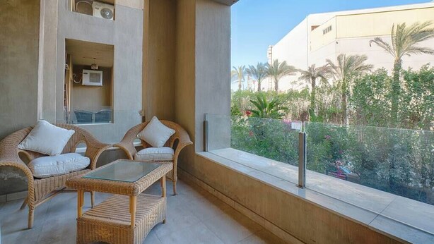 A balcony with wicker furniture, a glass table, and a view of palm trees and buildings. - The Suite, Palm Hills - V1811, Residential Suites near AUC by ELEGANT (New Cairo City)