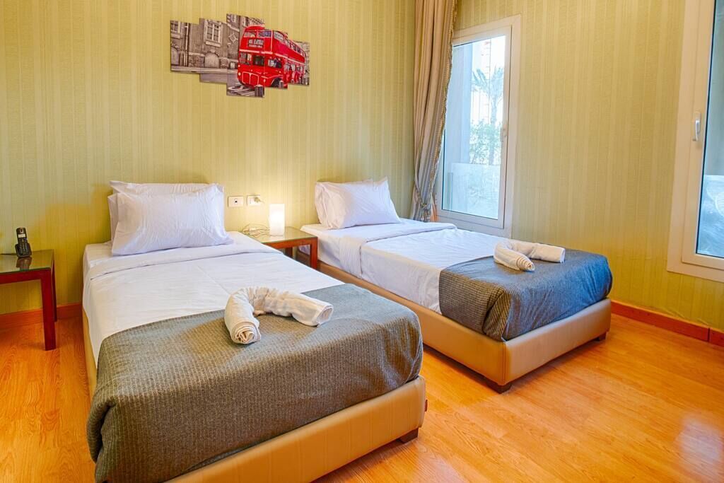 A hotel room with two single beds, a nightstand with a lamp, a window with curtains, and a red bus artwork on the wall.