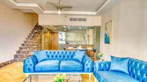 A living room with a blue sofa set, a glass coffee table, and a staircase leading to a kitchen. - The Lofts across AUC - C703, LOFTS BY ELEGANT - WITH POOL (New Cairo City)