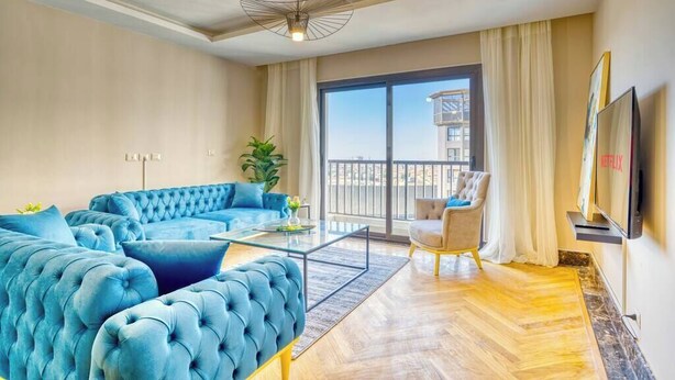 A modern living room with a blue sectional sofa, a glass coffee table, and a view of a building through large windows. - The Lofts across AUC - C703, LOFTS BY ELEGANT - WITH POOL (New Cairo City)