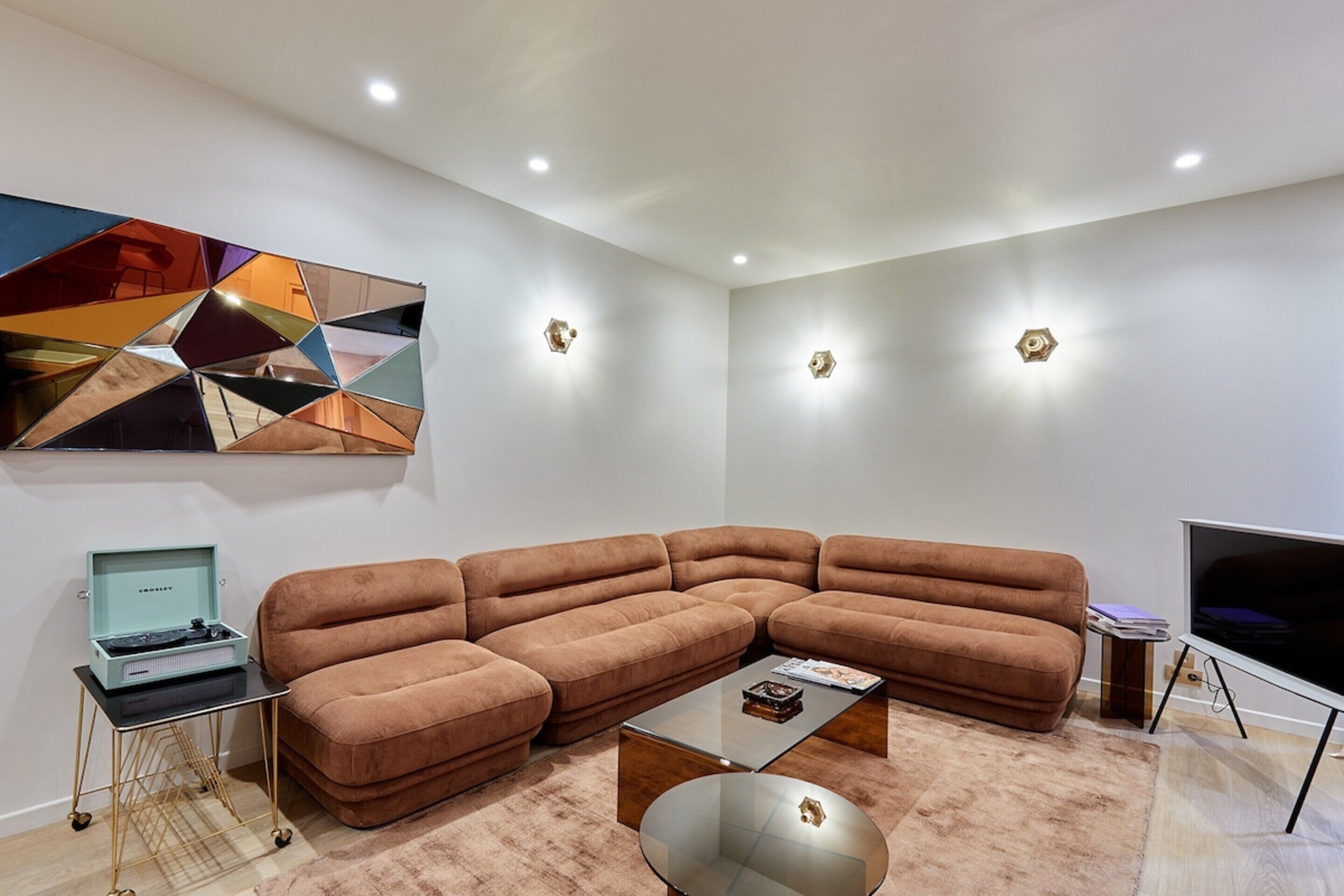 A modern living room with a sectional sofa, a glass coffee table, a flat-screen TV, and a geometric art piece on the wall.