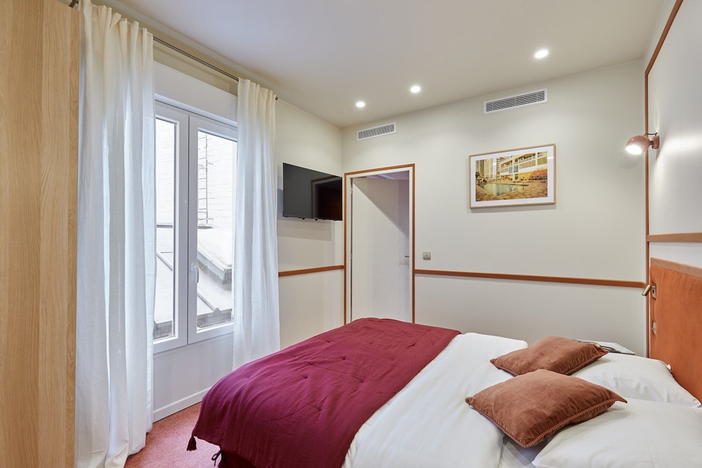Hotellihuone, jossa on sänky, seinälle asennettu televisio, taulu ja ikkuna, jossa on verhot.