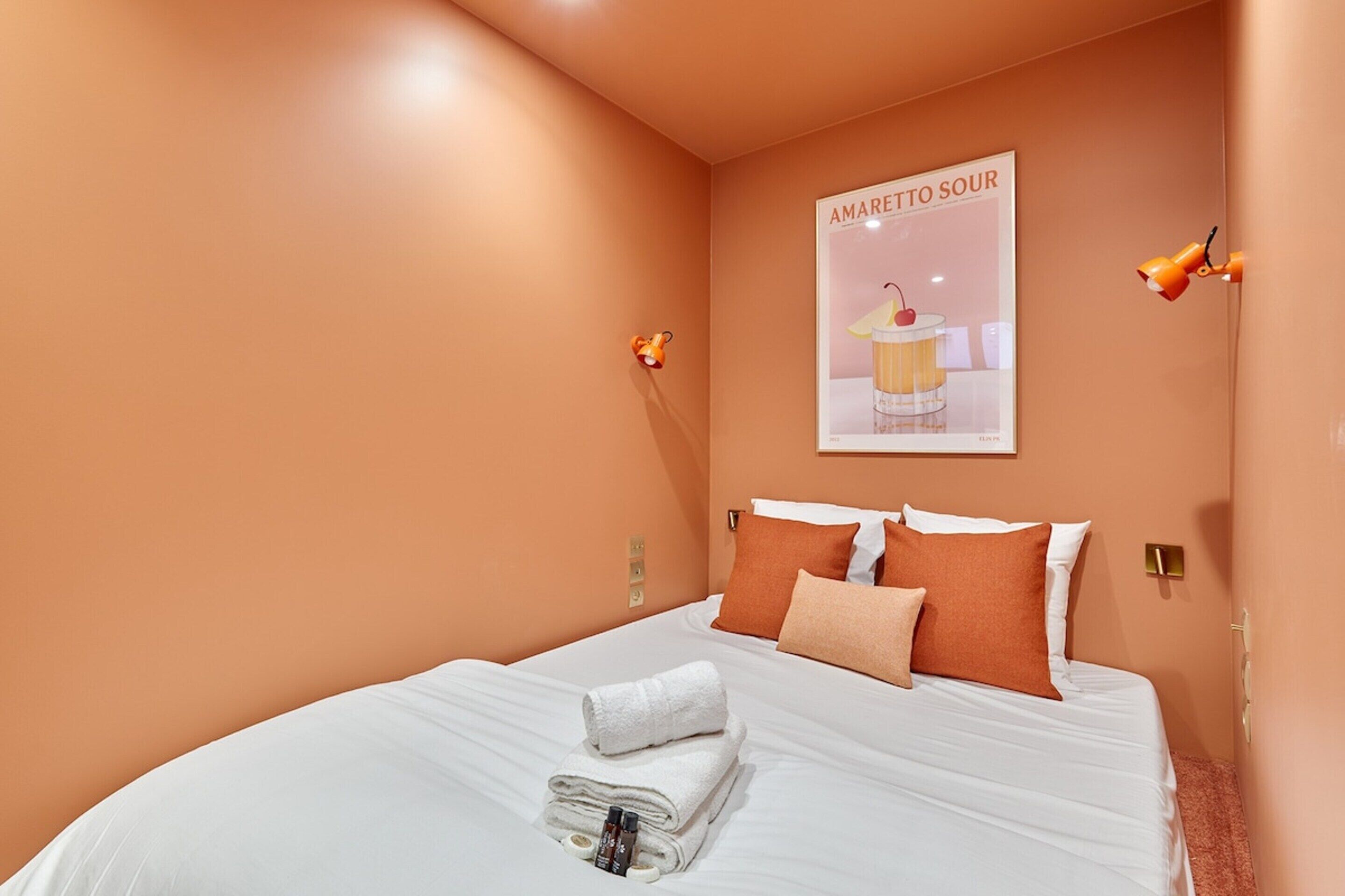 Een opgemaakt bed met wit en oranje beddengoed, een poster aan de muur en twee wandlampen.