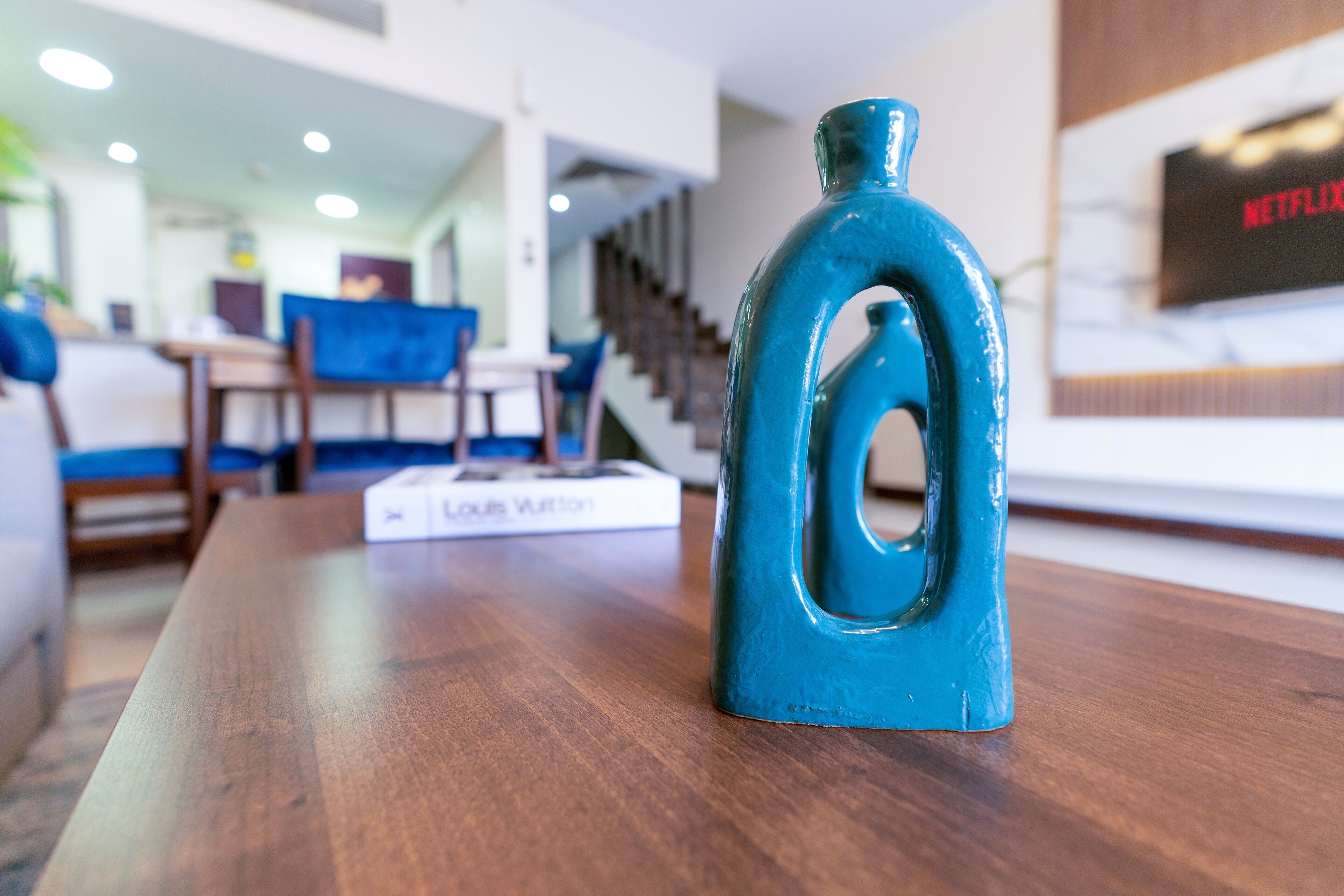A teal decorative bottle on a wooden table in a modern living room with a staircase and a TV displaying the Netflix logo.