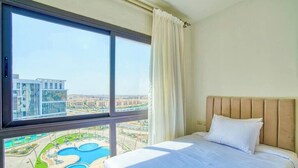 A hotel room with a large window, a bed with a headboard, and a view of a swimming pool and buildings. - The Lofts across AUC - A513, The Lofts by Elegant (New Cairo City)