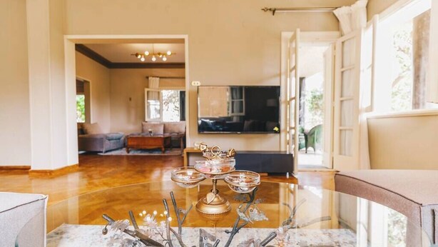 A dining table with a glass top and decorative metal stands, a chandelier, and a view of a living room with a sofa and television.