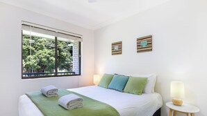 A hotel room with a bed, two bedside lamps, a window with blinds, and two wall art pieces. - Periwinkle - Ideal Beachfront Retreat Near Main Street with Ceiling Fans, Wi-Fi & Air Con (Sawtell)