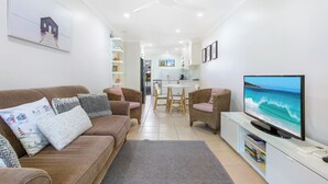 A modern living room with a sofa, chairs, a TV, and a kitchen in the background. - Periwinkle - Ideal Beachfront Retreat Near Main Street with Ceiling Fans, Wi-Fi & Air Con (Sawtell)