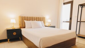 Una cama bien hecha con cabecera de madera, dos mesitas de noche con lámparas y una puerta a la derecha.