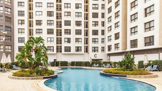 Una piscina rodeada de sillas de descanso y follaje, con un edificio alto al fondo.