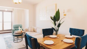 Un comedor con una mesa de madera dispuesta para cuatro, sillas tapizadas en azul, una planta en maceta y un sofá al fondo.