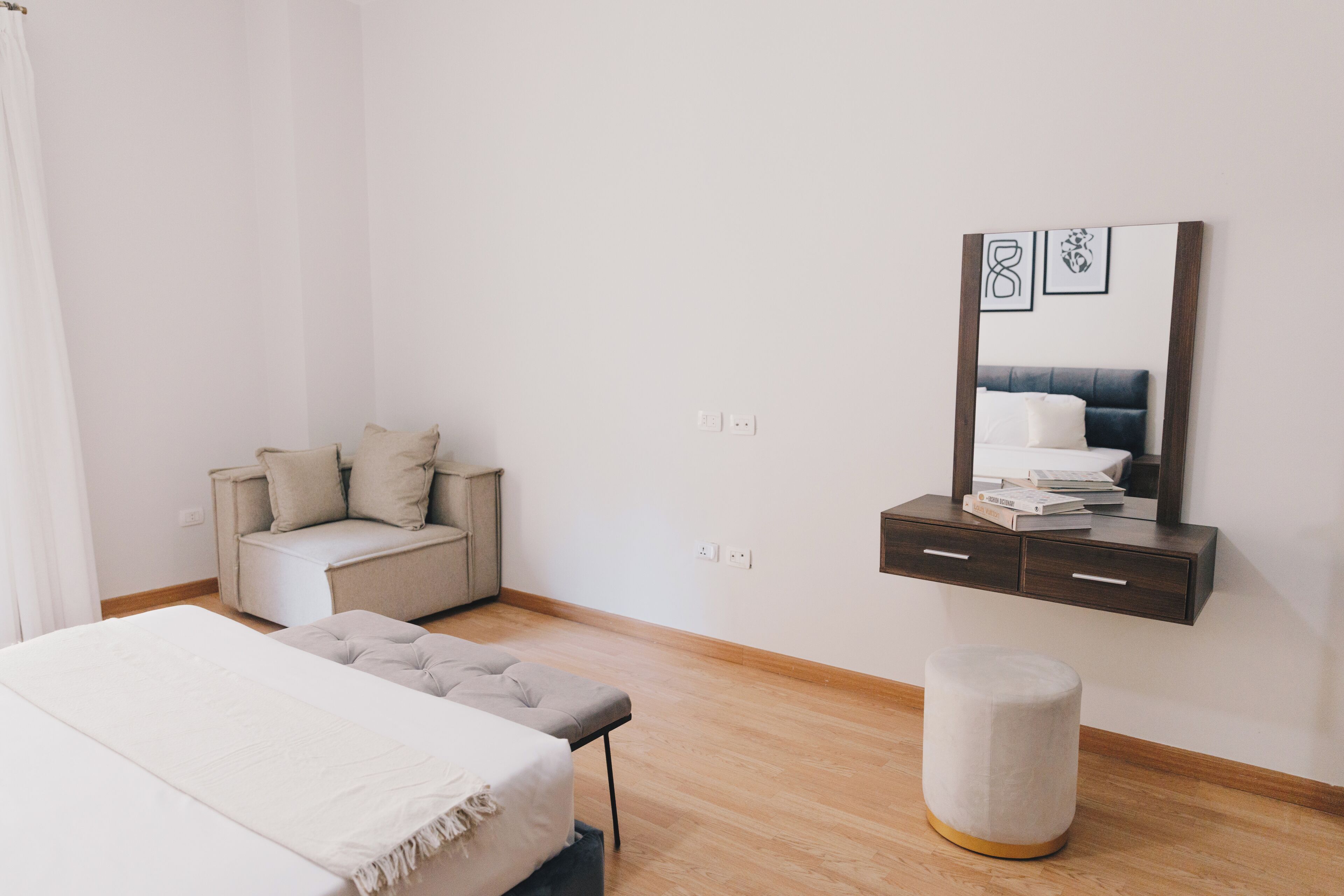 A bedroom with a bed, a bedside table, a chair, a small stool, and a wall-mounted cabinet with a mirror and framed pictures.