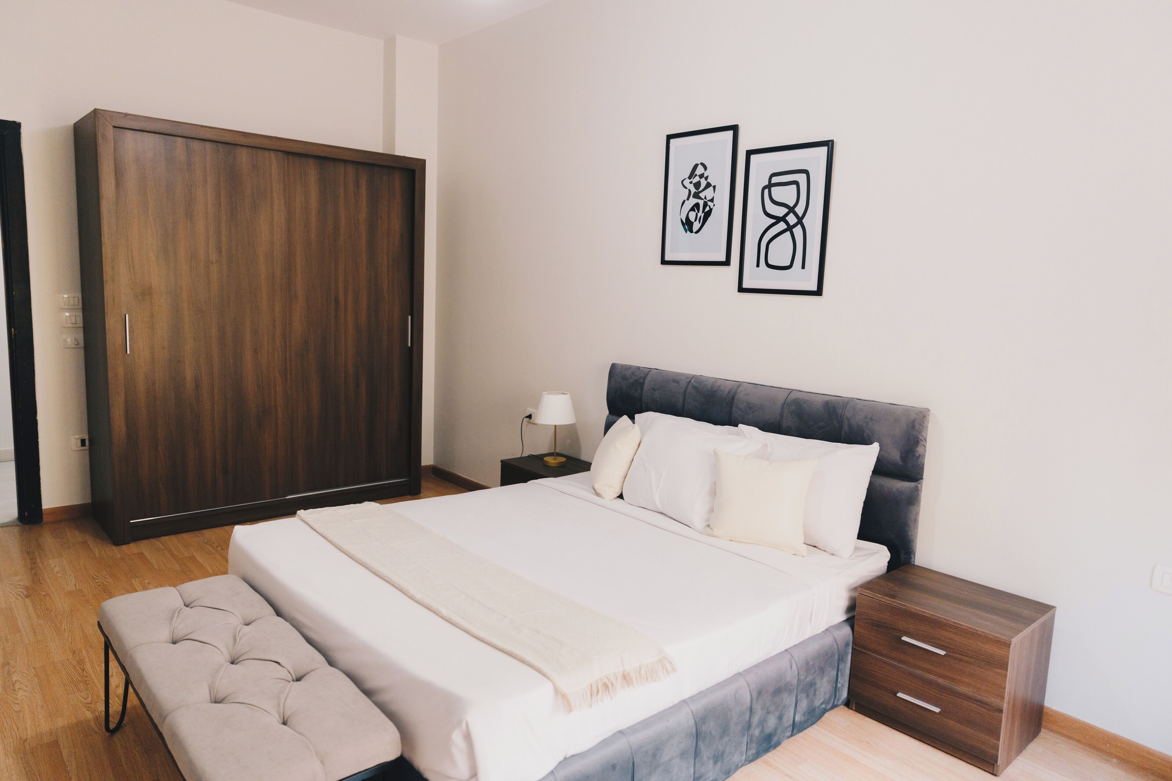 A bedroom with a large bed, a wardrobe, a bedside table with a lamp, and two framed pictures on the wall.