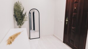 A room with a white bed, a mirror, a potted plant, and a wooden door.